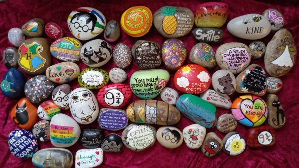 A collection of painted rocks with various designs, quotes, and drawings, arranged on a red textured background.
