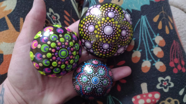 A hand holding three colourful mandala stones with intricate dot patterns in vibrant greens, purples, and blues, against a patterned fabric background.