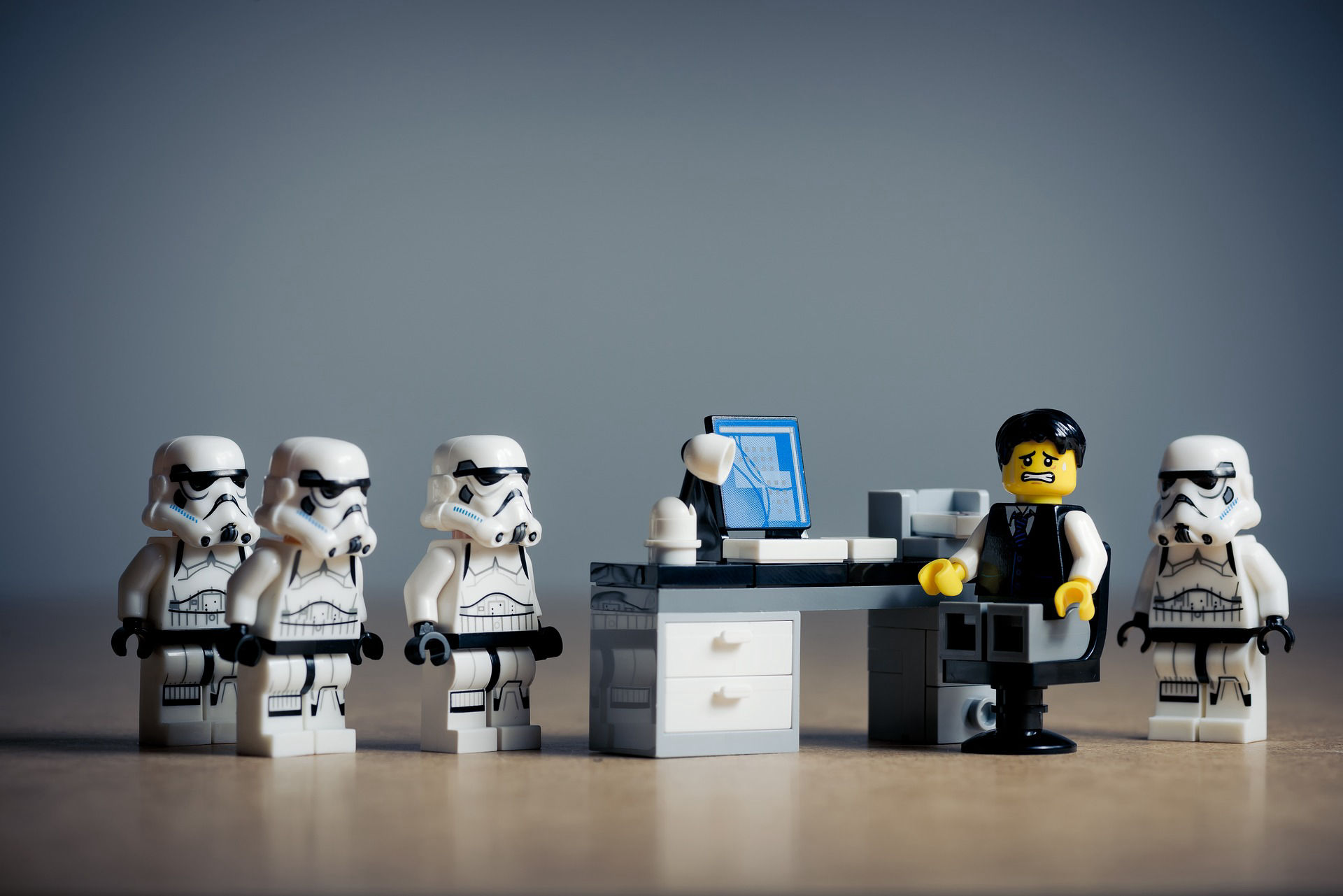 LEGO-style minifigures in an office scene: four stormtroopers line up in front of a desk with a computer, while an anxious-looking figure sits behind it.