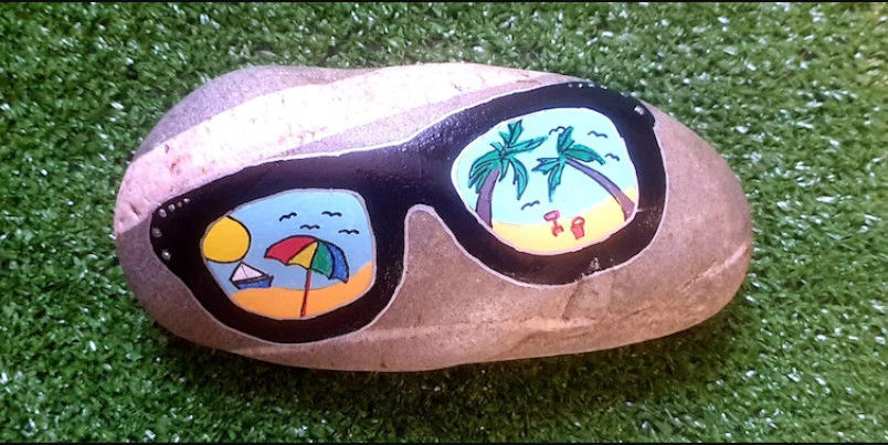 A painted rock features sunglasses with beach scenes reflecting palm trees, a sun umbrella, and birds. It rests on vibrant green grass, conveying a summery vibe.