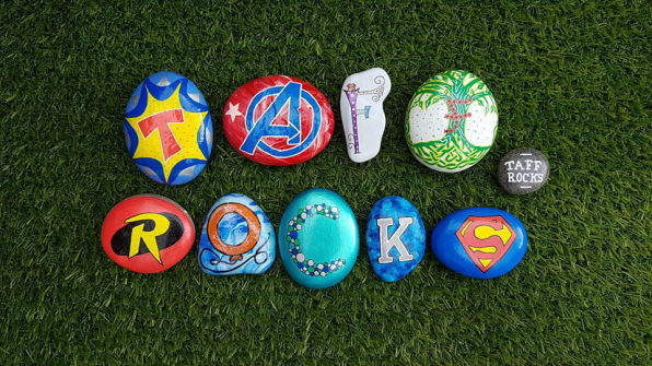 Colourful painted rocks on grass spell "TAFF ROCKS." Each rock features superhero symbols or nature designs, creating a fun and vibrant scene. Created by worldwide friends of Taff Rocka