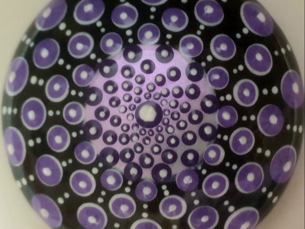 A round, black object displays a mesmerizing mandala pattern of concentric circles in shades of purple and white, creating a hypnotic and vibrant design.