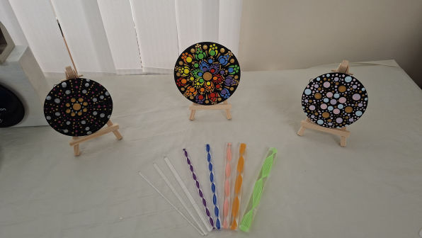 Three colourful dot mandala paintings on small easels are displayed on a table. In front, there are eight, multi-coloured dotting tools.