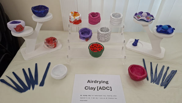 Colourful handmade clay bowls are displayed on stands and a clear rack, accompanied by blue sculpting tools. A sign reads "Airdrying Clay [ADC]."