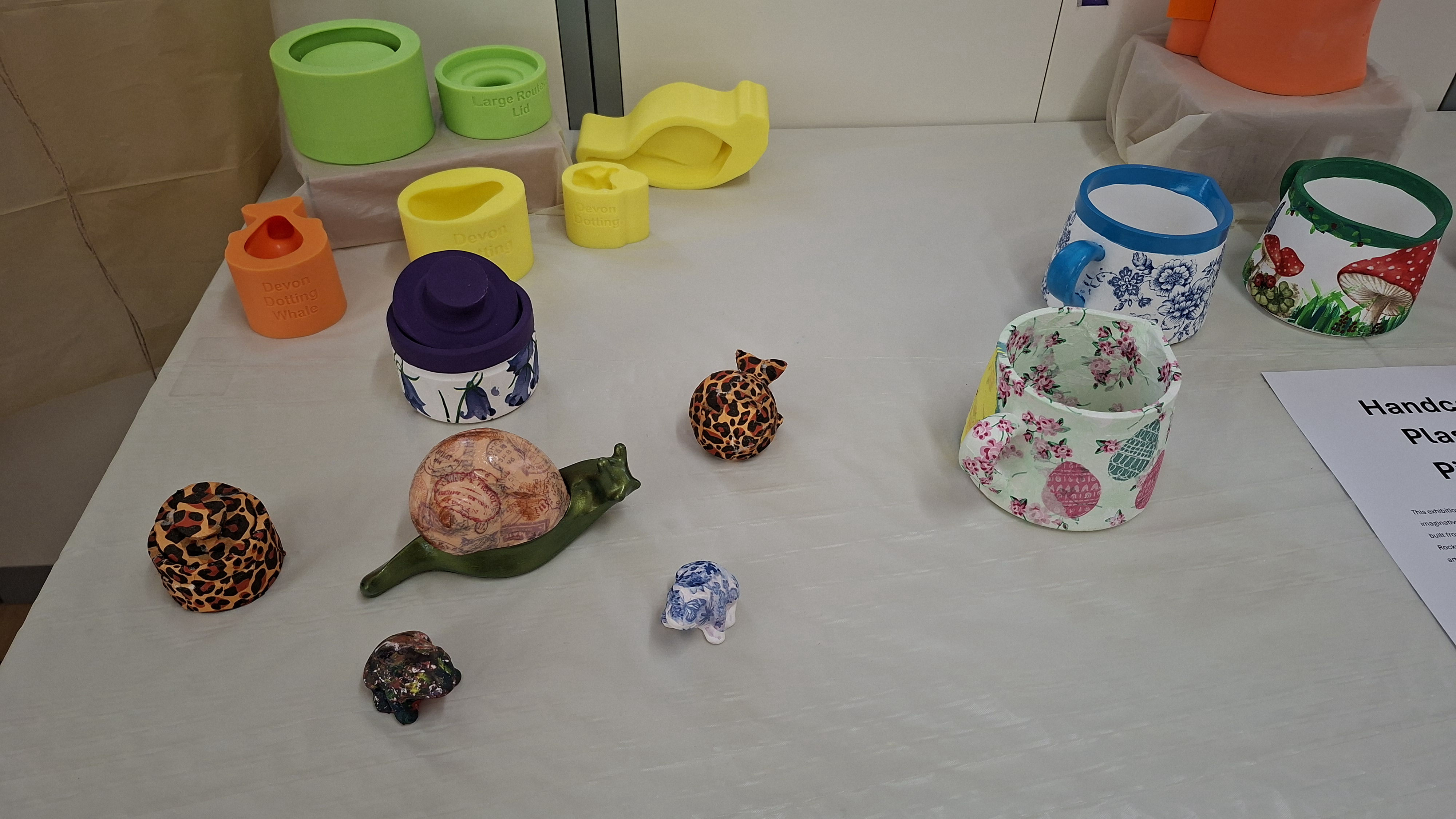 A display of colourful, handmade jugs and decorative animal figurines on a white tablecloth. Items include floral and patterned designs, radiating creativity.