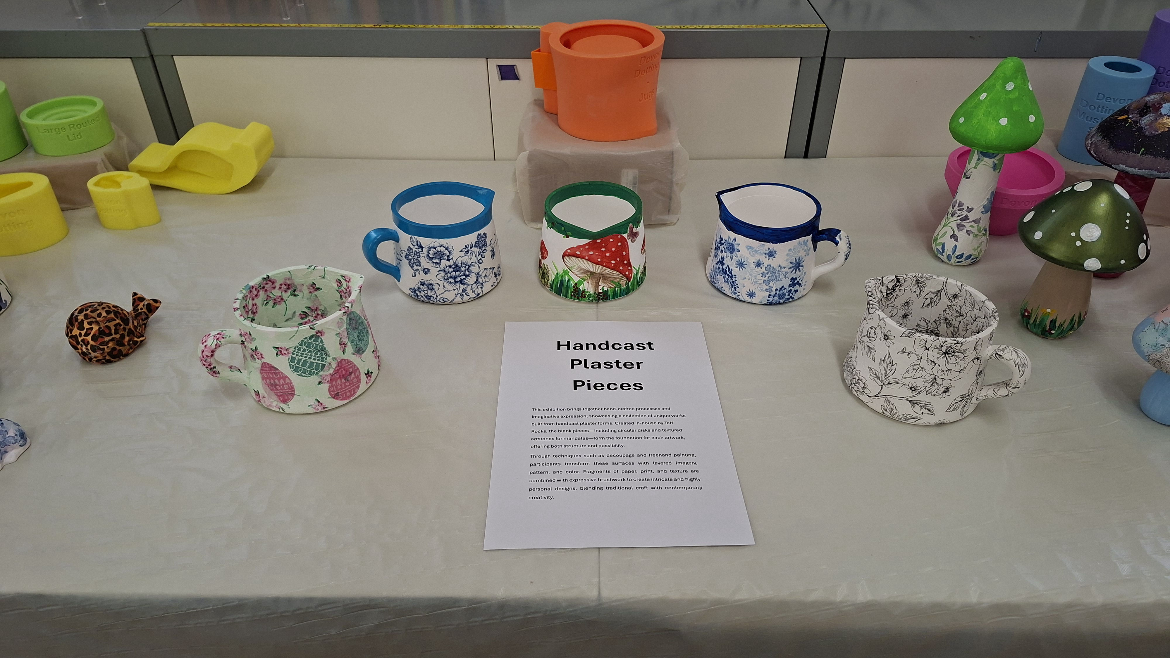Handcrafted plaster jugs with colourful designs are displayed on a table. A sign titled "Handcast Plaster Pieces" is placed in the centre.