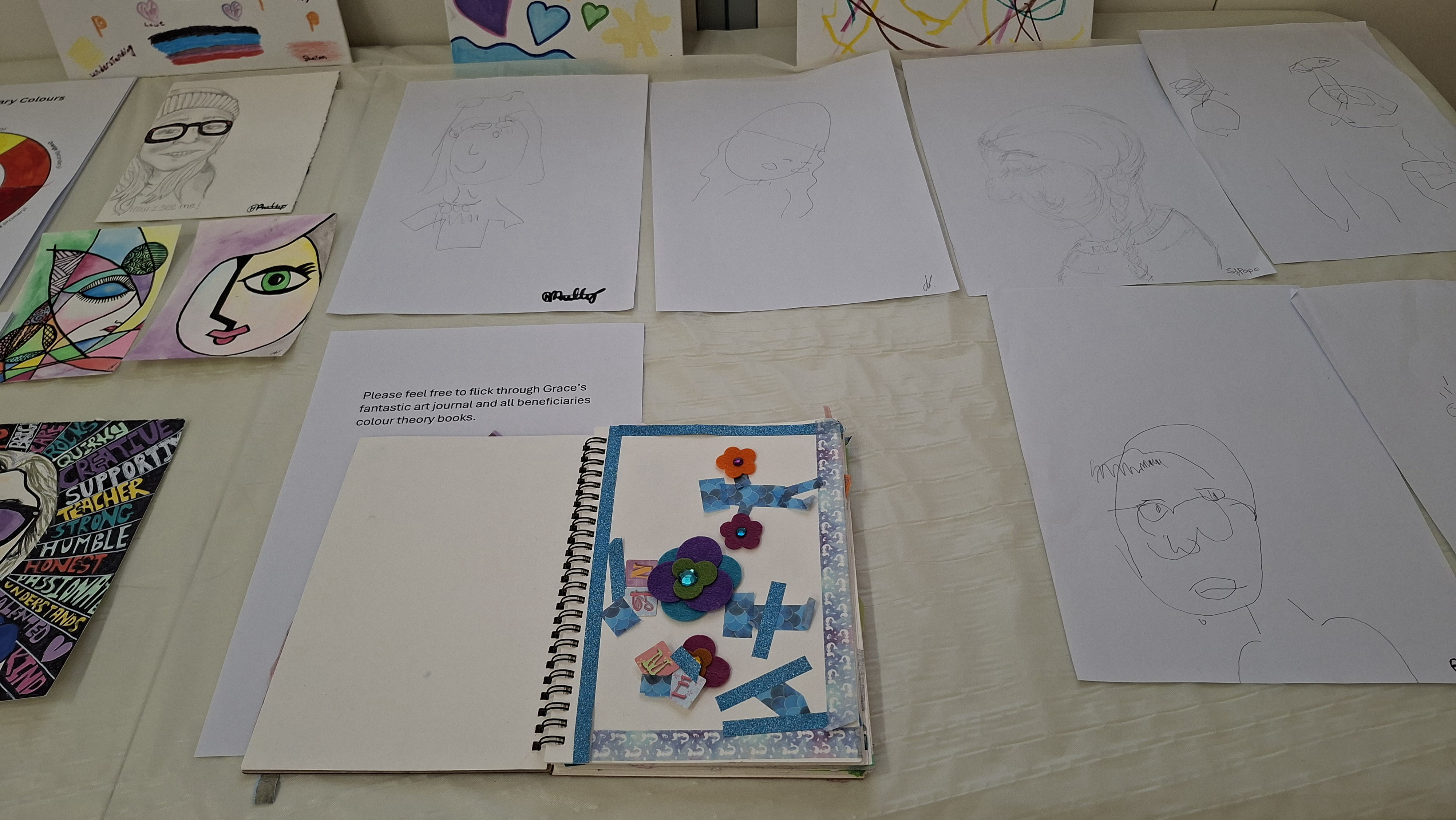 Art display with sketches and colourful abstract faces. A decorated journal features layered paper designs and fabric flowers. Creative and vibrant.
