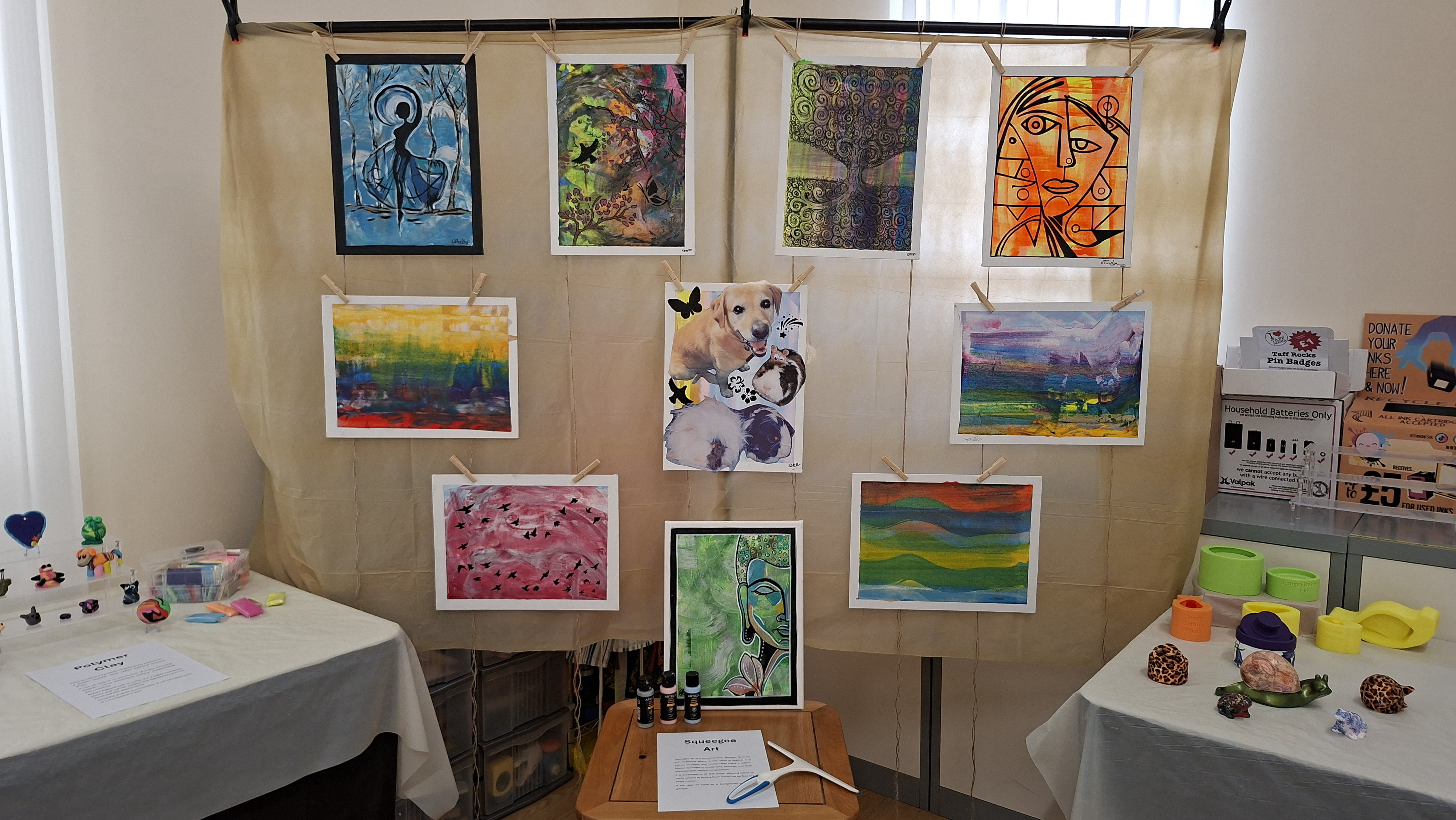 Art display featuring nine colourful paintings hung on a beige canvas backdrop. Tables on either side hold small crafts and informational papers. Bright and inviting.