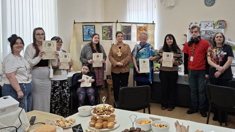 Our group of artists in Smyrna holding certificates, with their colourful artwork behind them. A table with snacks is in the foreground. Smiles convey a celebratory mood.