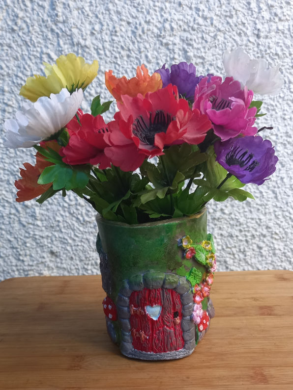 Vibrant artificial flowers in a whimsical green air dried clay vase with a red door and mushrooms, set against a textured white wall, evoke a playful garden charm.