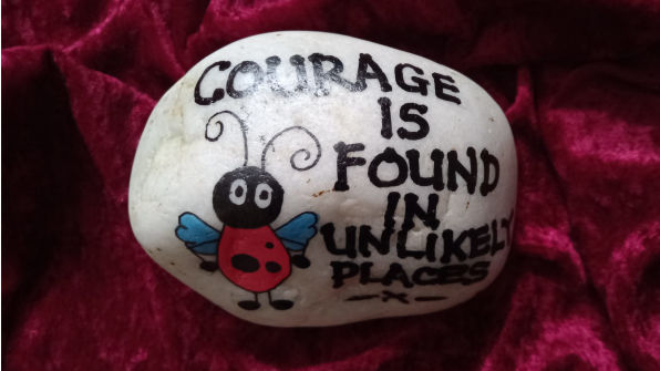 A painted rock with a ladybug drawing and the words courage is found in unlikely places on a red velvet background, conveying a message of inspiration.
