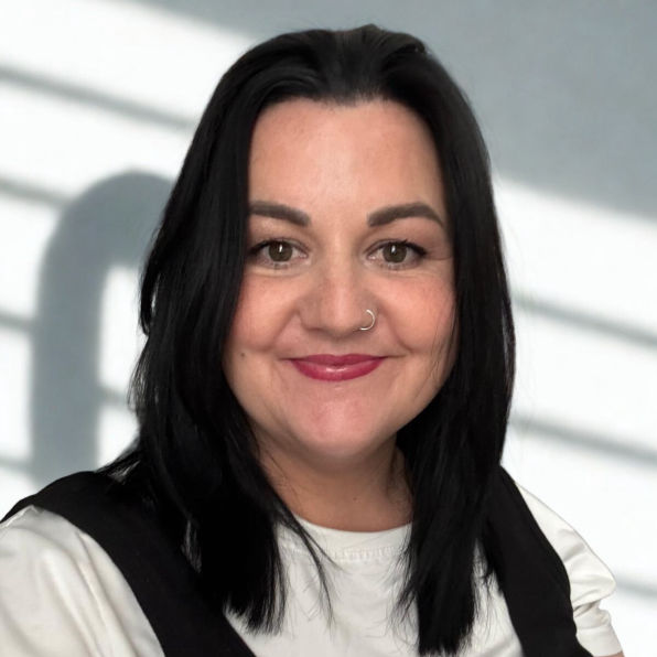 An image of Hayley, smiling and wearing a white top under a black vest. The background features soft shadows on a light wall, creating a calm tone.