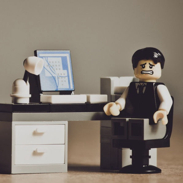 A worried LEGO-style figure sits at a desk with a computer and lamp. The scene conveys stress, with muted colours enhancing the sombre office setting.